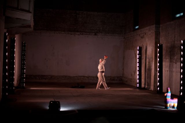 Performance How Happy We Would Be 2011 by Corali Dance Company featuring dancer Bethan Kendrick who holds her arm up as she walks towards a rainbow coloured pinata on the concrete stage of Dilston Grove, Café Gallery Projects